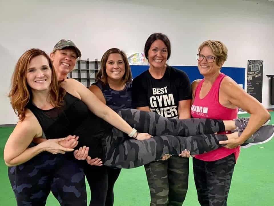 Group of strong, smiling women in athletic wear lifting one of their teammates at Results by Design Fitness